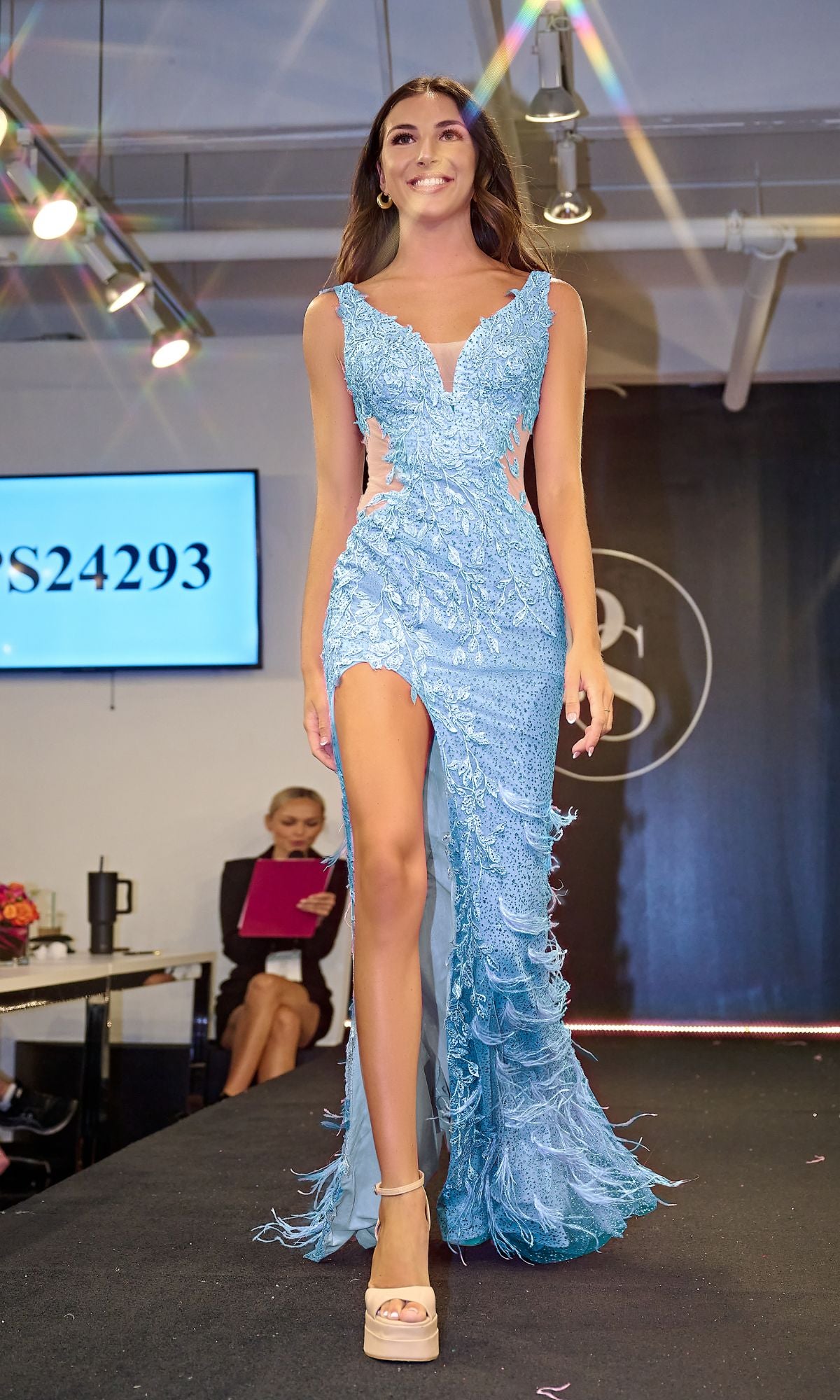 A woman walks the runway in the Long Prom Dress PS24293 by Portia and Scarlett, a light blue gown featuring lace embroidery and a thigh-high slit; in the background are a seated woman and a digital display.